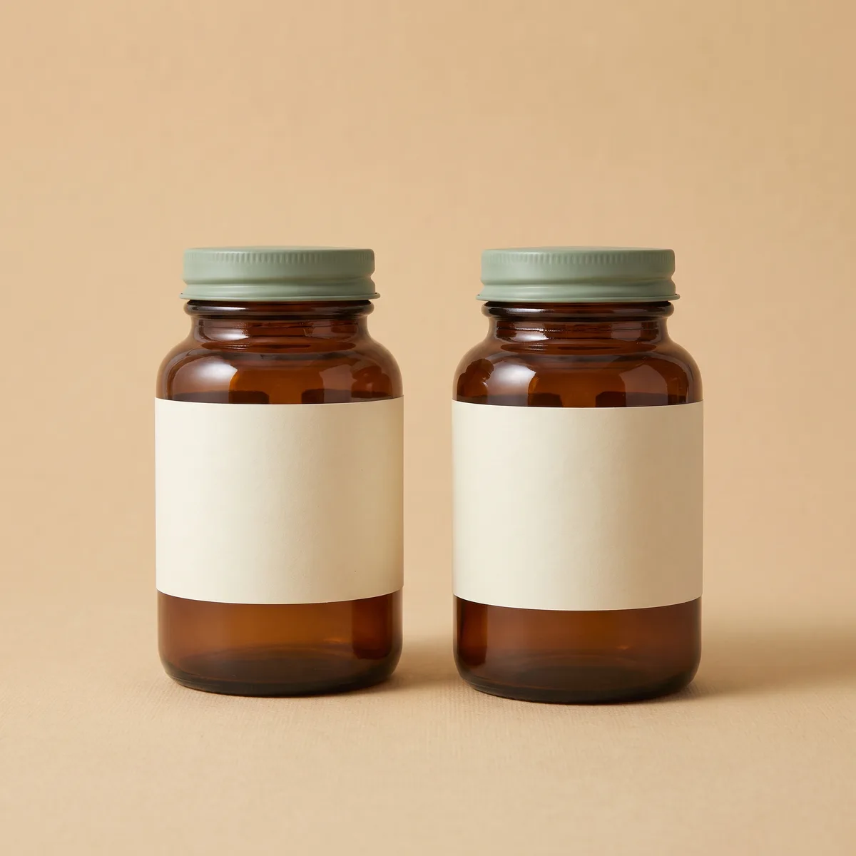 Two amber jars with sage lids standing side by side on an oat backdrop